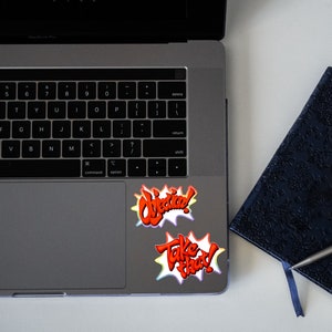 May include: A laptop computer with two colorful stickers on the trackpad. The stickers say "Objective!" and "Take That!" in a comic book style font.