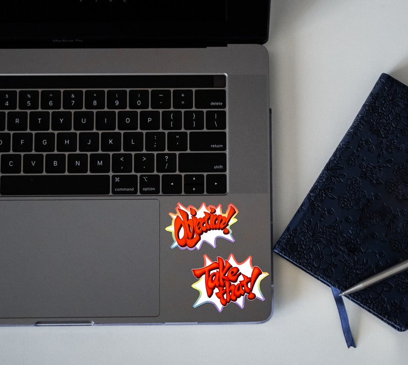 May include: A laptop computer with two colorful stickers on the trackpad. The stickers say "Objective!" and "Take That!" in a comic book style font.