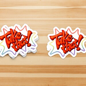 May include: Two colorful stickers with the text "Take that!" in a red, white, and blue color scheme. The stickers are on a wooden surface.