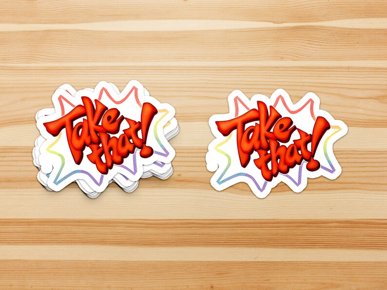 May include: Two colorful stickers with the text "Take that!" in a red, white, and blue color scheme. The stickers are on a wooden surface.