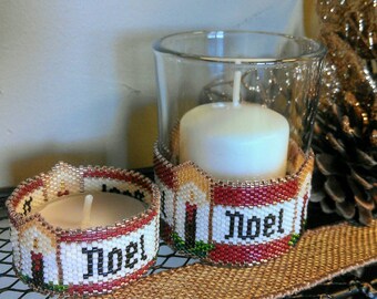 May include: Two beaded candle holders with the word "Noel" in black lettering. The candle holders are red, white, and gold. The candle holders are on a brown surface with a pine cone.