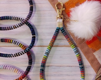 May include: A colorful beaded keychain with a gold clasp and a white fluffy pom-pom. The keychain is made of small, colorful beads in a geometric pattern. The keychain is a great way to add a pop of color to your keys or bag.