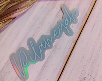May include: A holographic sticker with the word "Pilgrim" written in a cursive font. The sticker is on a white wooden surface.