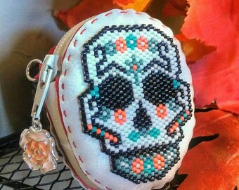 May include: A white leather coin purse with a beaded skull design. The skull is decorated with black, orange, and turquoise beads. The purse has a zipper closure and a small rose charm attached to the zipper pull.