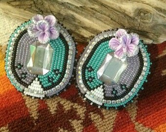 May include: A pair of beaded earrings with a turquoise and purple color scheme. Each earring features a large, clear crystal in the center and a purple flower embellishment.