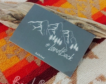 May include: A gray sticker with a white outline of mountains and trees. The text "#LandBack" is written in white cursive lettering. The text "Bead Be Charmed" is written in white at the bottom of the sticker.