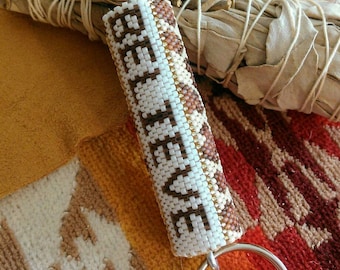 May include: A white beaded keychain with brown and gold accents. The keychain is shaped like a cylinder and has the word "Believe" beaded on it. The keychain is attached to a silver key ring.