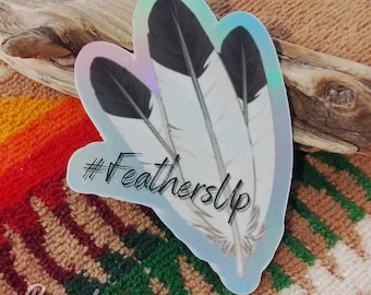 May include: A holographic sticker with a design of three white and black feathers. The sticker has the text "#FeathersUp" in black lettering.