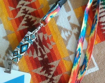 May include: A colorful lanyard with a beaded keychain attached to a white badge with the word "ATE" on it. The lanyard is on a patterned fabric background.