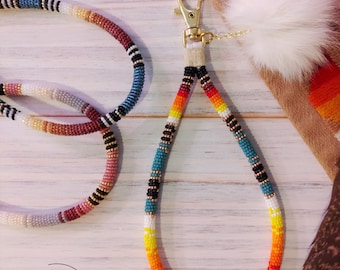 May include: Two beaded lanyards with a gold key chain and a white fur pom-pom. The lanyards are made with colorful beads in a variety of patterns. The text "Bead Charmed Design Studio Est'd 2001" is visible in the lower left corner of the image.