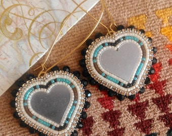 May include: A pair of heart-shaped earrings with a mirror in the center. The earrings are beaded with black, white, and turquoise beads. The earrings are hanging from gold hoops.