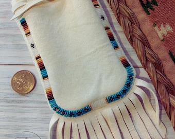 May include: A white leather pouch with a beaded trim and fringe. The pouch is decorated with turquoise, brown, and white beads. The pouch is sitting on a wooden surface.
