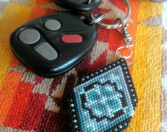 May include: A black car key with three buttons and a red button is attached to a beaded keychain with a diamond-shaped design in shades of blue, white, and black.
