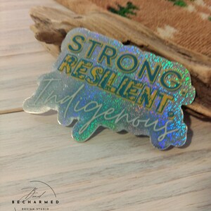 May include: A holographic sticker with the words "STRONG RESILIENT Indigenous" in teal and yellow. The sticker has a glittery, iridescent finish and is on a light wood surface.
