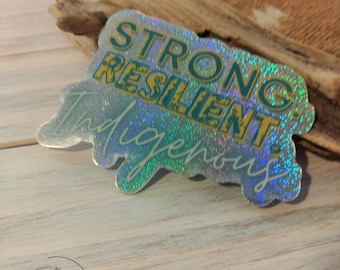 May include: A holographic sticker with the words "STRONG RESILIENT Indigenous" in teal and yellow. The sticker has a glittery, iridescent finish and is on a light wood surface.