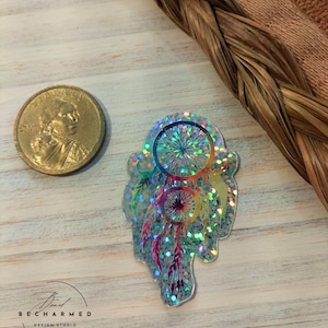 May include: A holographic iridescent dreamcatcher sticker with a rainbow color scheme. The sticker is on a light wood background.