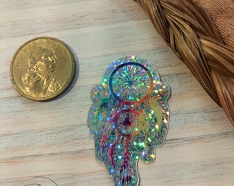 May include: A holographic iridescent dreamcatcher sticker with a rainbow color scheme. The sticker is on a light wood background.