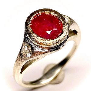May include: A silver ring with a red ruby gemstone set in a gold bezel. The ring has two smaller diamonds on the band.
