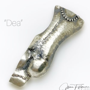 May include: A silver-toned, abstract sculpture of a female torso with a beaded necklace detail. The sculpture is titled "Dea".