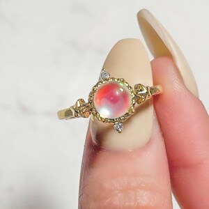 May include: A gold ring with a round, iridescent gemstone surrounded by a delicate gold filigree design. The ring is set with small, clear gemstones.