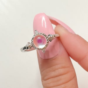May include: A silver ring with a round iridescent pink gemstone set in a delicate silver frame. The ring is adorned with small silver accents.