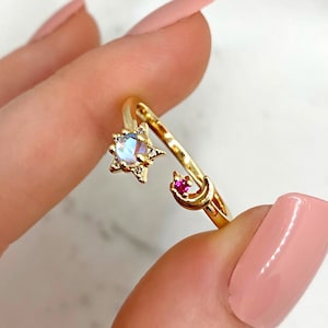 May include: Gold ring with a moon and star design. The star has a clear iridescent stone in the center and the moon has a small pink stone.