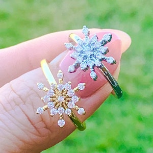 May include: Two gold rings with a snowflake design set with clear cubic zirconia stones. The rings are on a finger.