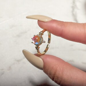 May include: A gold ring with a round iridescent gemstone surrounded by a decorative setting. The ring is on a finger.
