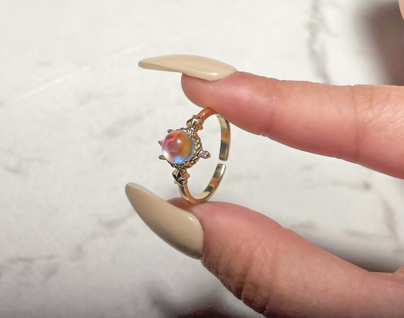 May include: A gold ring with a round iridescent gemstone surrounded by a decorative setting. The ring is on a finger.