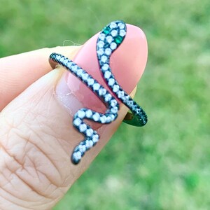 May include: A black snake ring with clear and green stones. The snake is coiled and the head is facing the right side of the image.