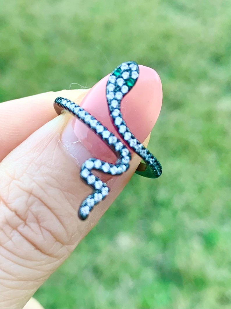 May include: A black snake ring with clear and green stones. The snake is coiled and the head is facing the right side of the image.