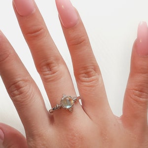 May include: A silver ring with a large oval-shaped iridescent gemstone surrounded by smaller clear gemstones. The ring is on a person's finger.