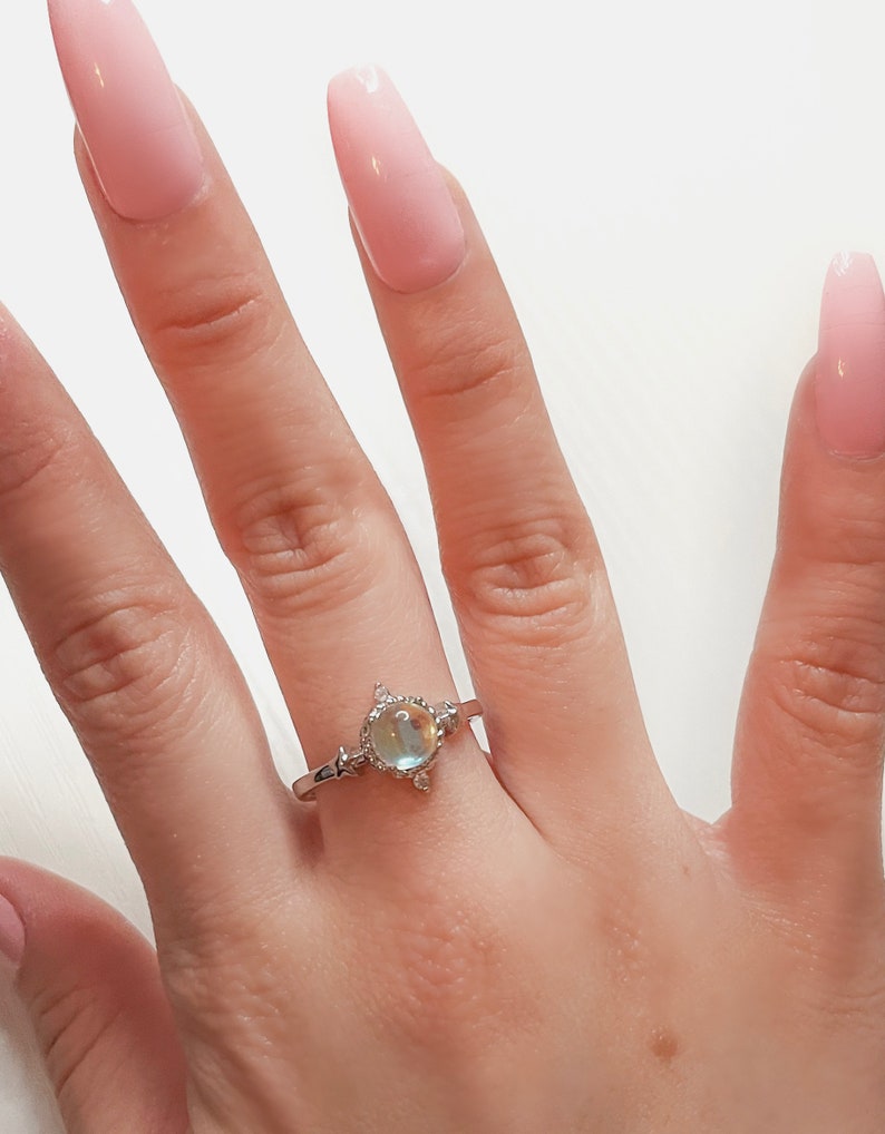 May include: A silver ring with a large oval-shaped iridescent gemstone surrounded by smaller clear gemstones. The ring is on a person's finger.