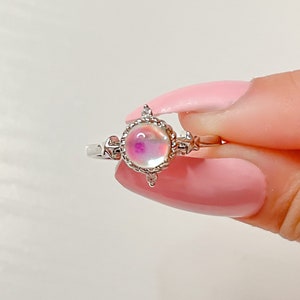 May include: A silver ring with a round iridescent opal-like stone set in a decorative silver bezel. The ring has a delicate design with small accents.