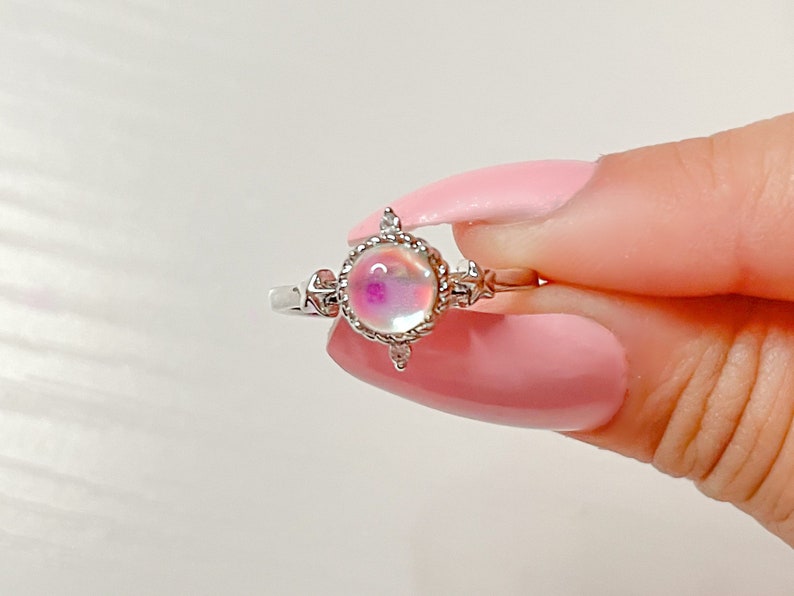 May include: A silver ring with a round iridescent opal-like stone set in a decorative silver bezel. The ring has a delicate design with small accents.