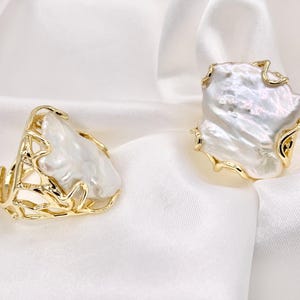 May include: Two rings featuring large, irregular white baroque pearls set in gold-toned metal bands. The metalwork has an open, branch-like or coral-inspired lattice design that frames the lustrous, organic shape of each pearl.