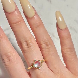 May include: A gold ring with a large oval-shaped gemstone set in a decorative frame. The gemstone is a pale pink and white color with a pearlescent finish. The ring is on a finger with light beige nail polish.