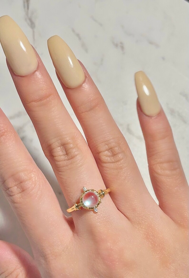 May include: A gold ring with a large oval-shaped gemstone set in a decorative frame. The gemstone is a pale pink and white color with a pearlescent finish. The ring is on a finger with light beige nail polish.