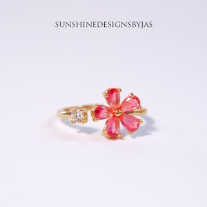 May include: A gold ring with a pink flower design. The flower has six petals and a small clear stone in the center. The ring is adjustable and can be worn on any finger.