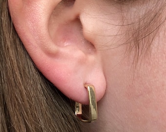 May include: Close-up of a gold-colored square hoop earring. The earring is a simple, geometric design with a polished finish. The earring is worn on an ear, with the skin tone visible in the background.
