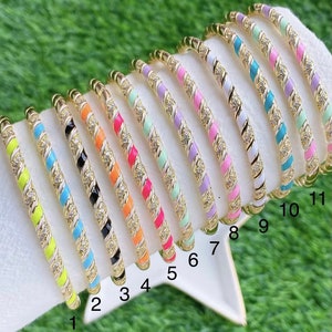 May include: A collection of gold-toned bangles with colorful enamel stripes and sparkling accents. Each bangle features a different color combination, including yellow, blue, black, orange, pink, and purple. The bracelets are displayed on a white surface against a green background.