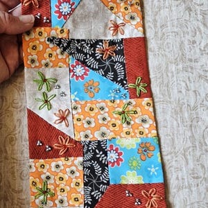 Può includere: Tessuto patchwork colorato con motivi floreali arancioni, blu e neri. Il tessuto è impreziosito da fiori ricamati e piccole perline d'argento. Il design combina forme geometriche e motivi floreali, creando un aspetto vibrante e artigianale.