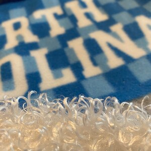 May include: Close-up of a white, curly, textured fabric in the foreground. In the background, a blue and white checkered pattern with the text "NORTH CAROLINA" is visible, suggesting a collegiate theme.