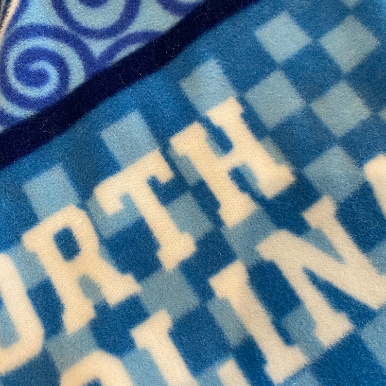 May include: Close-up of a blue and white fleece blanket. The blanket features the word "NORTH CAROLINA" in large white letters. The background has a checkered pattern in shades of blue and a swirl design.