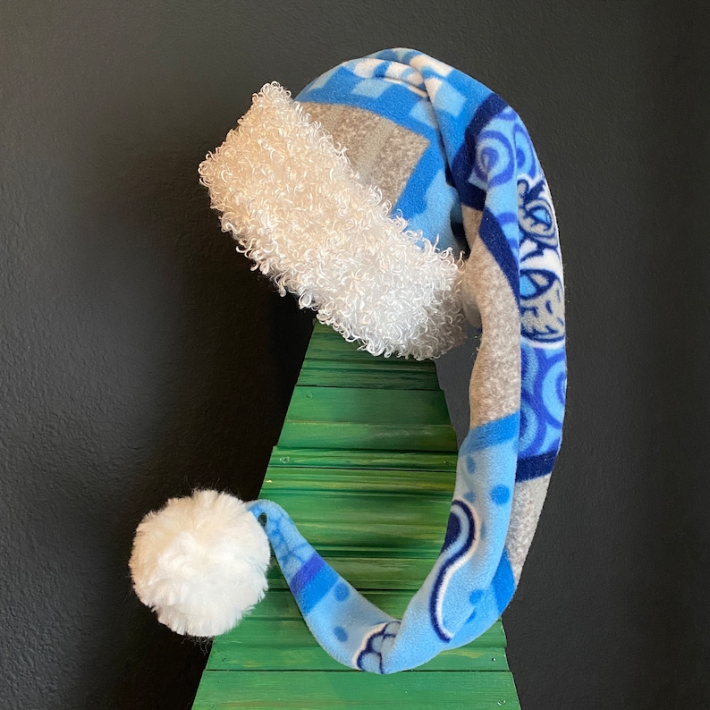 May include: A whimsical Santa hat with a long, pointed shape. The hat features a blue and gray patterned fabric with white faux fur trim and a fluffy white pom-pom. The hat is displayed on a green wooden Christmas tree.