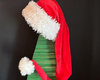 Può includere: Un cappello rosso di Babbo Natale con finiture bianche e soffici e un pompon bianco all'estremità. Il cappello è drappeggiato su un albero di Natale di legno verde. Lo sfondo è nero.