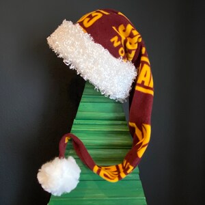 May include: A maroon and gold Santa hat with a white fluffy trim and a matching pom-pom. The hat is made of a soft fabric and features a repeating pattern. The hat is displayed on a green wooden Christmas tree.
