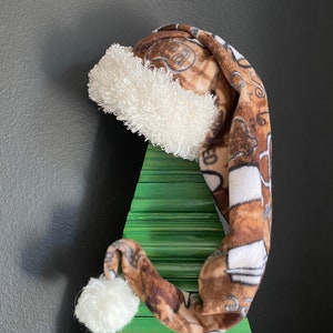 May include: A brown and white patterned Santa hat with a white fluffy trim and a white pom-pom. The hat is resting on a green wooden Christmas tree.