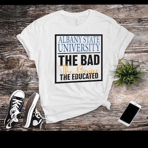 May include: White t-shirt with the text "ALBANY STATE UNIVERSITY" in blue, "THE BAD" in black, "The Boujee" in gold, and "THE EDUCATED" in black. Black and white sneakers and a smartphone are also visible.