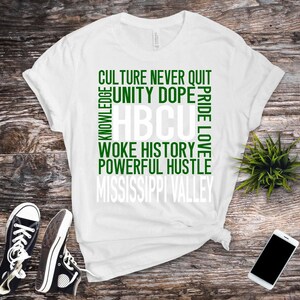 May include: White t-shirt with green and white text. The text includes the words "Culture Never Quit", "Unity Dope", "HBCU", "Woke History", "Powerful Hustle", and "Mississippi Valley". Black sneakers and a smartphone are also in the image.